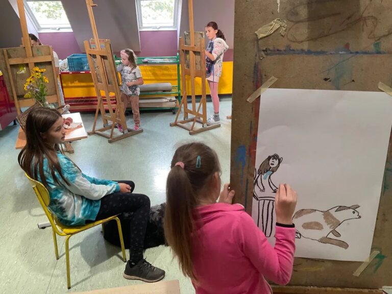Děti malují u stojanů v ateliéru, uprostřed dívka v modrém se psem