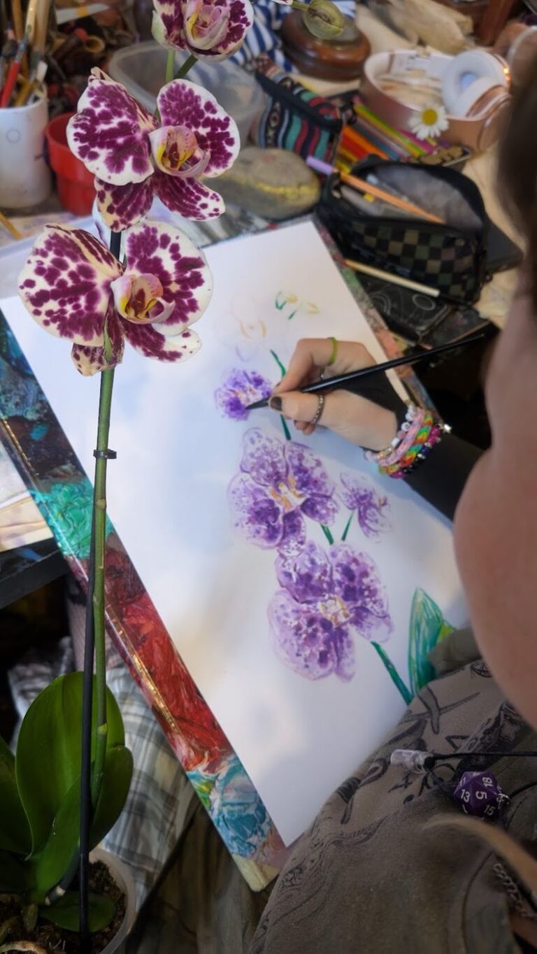 Studentka maluje akvarelem fialové orchideje podle živé rostliny
